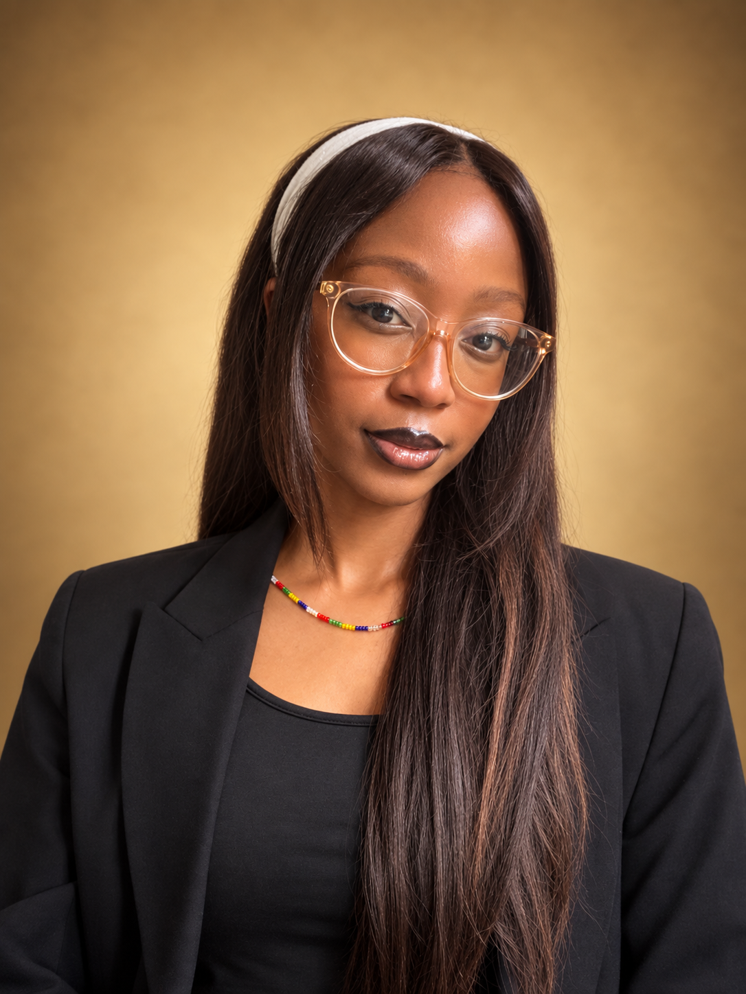 Professional headshot of a woman with long dark hair, clear glasses, a white headband, black blazer, and a colorful beaded necklace against a gold background.